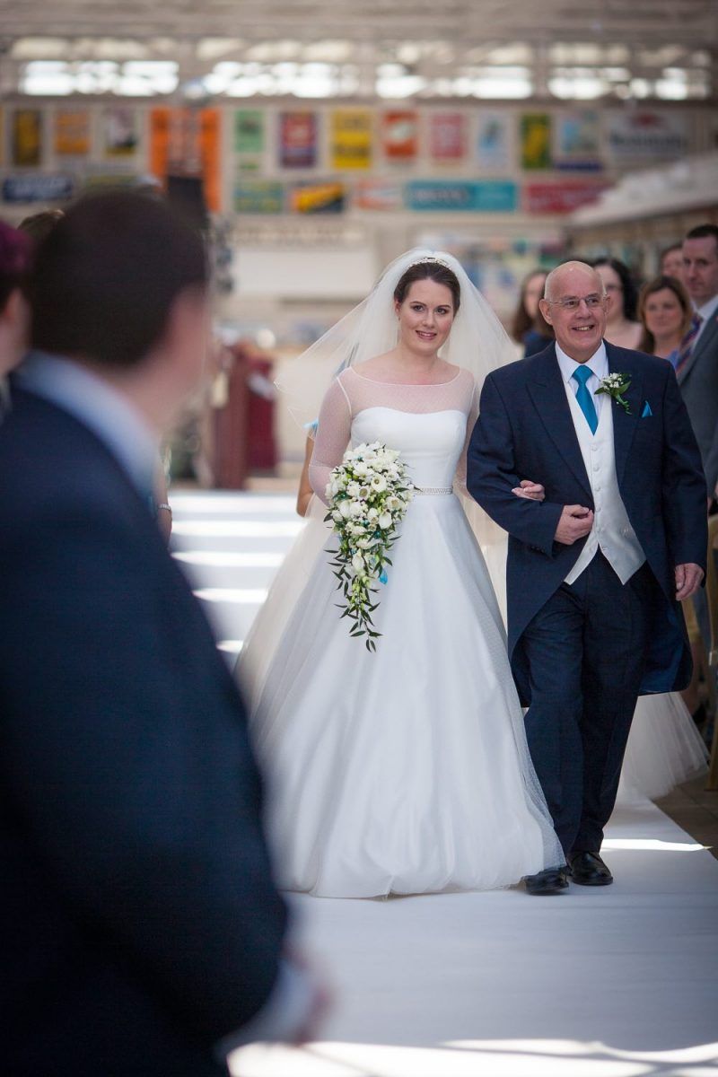 Emma walks down the aisle with her Dad against a backdrop of vintage signage at their Buckinghamshire Railway Centre wedding