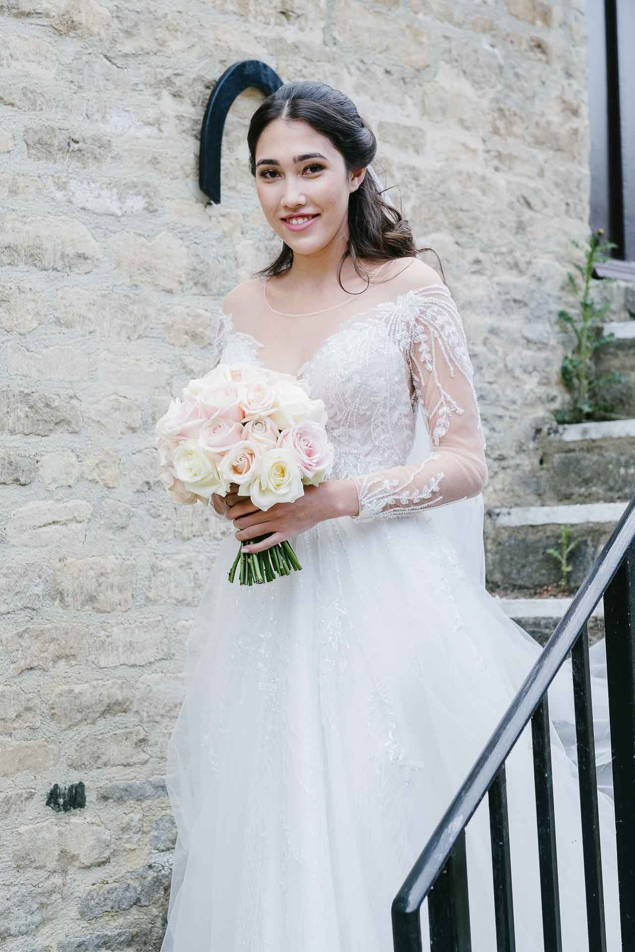 Nadia holding her bouquet in her wedding dress on the steps at Minster Mill. Photography by Des Dubber. Videography by Veiled Productions - fun wedding films