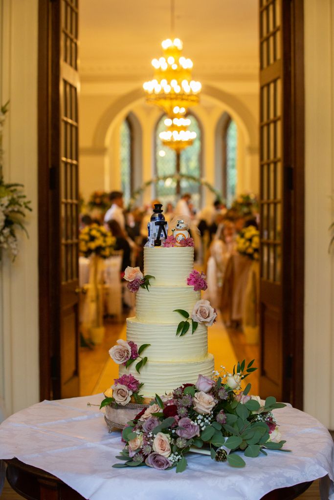 Incredible 4 tier wedding cake with ivory coloured frosting and fresh flowers decorating it. A nod to Star Wars with two robot bride and groom cake topper characters on the top tier. Photo thanks to Mark Lord Photography.