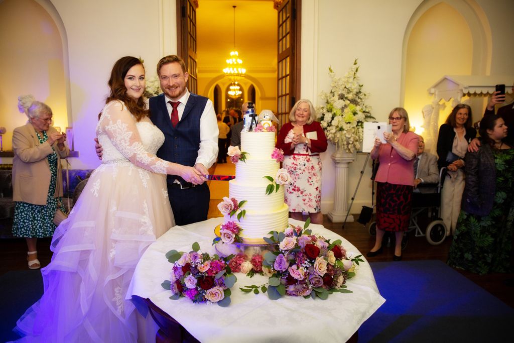 Aimee and Simon cutting their 4 tier wedding cake surrounded by their guests at Clearwell Castle. Photo thanks to Mark Lord Photography.