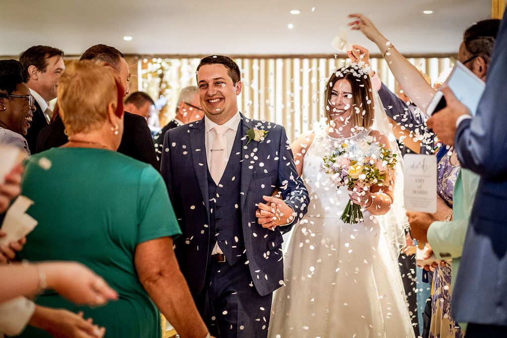 Amy and Mario exit as newlyweds whilst guests throw confetti. Wedding photo by Allister Freeman. Videography by Veiled Productions - unique wedding videographer Bury Court Barn
