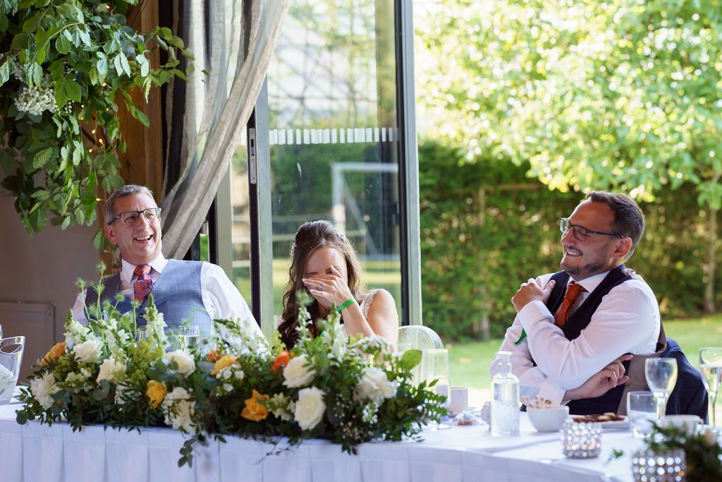 Bex and Dan laughing at the best man speech in front of the open glass doors at Redhouse Barn.. Photo courtesy of J Bidmead Photography.