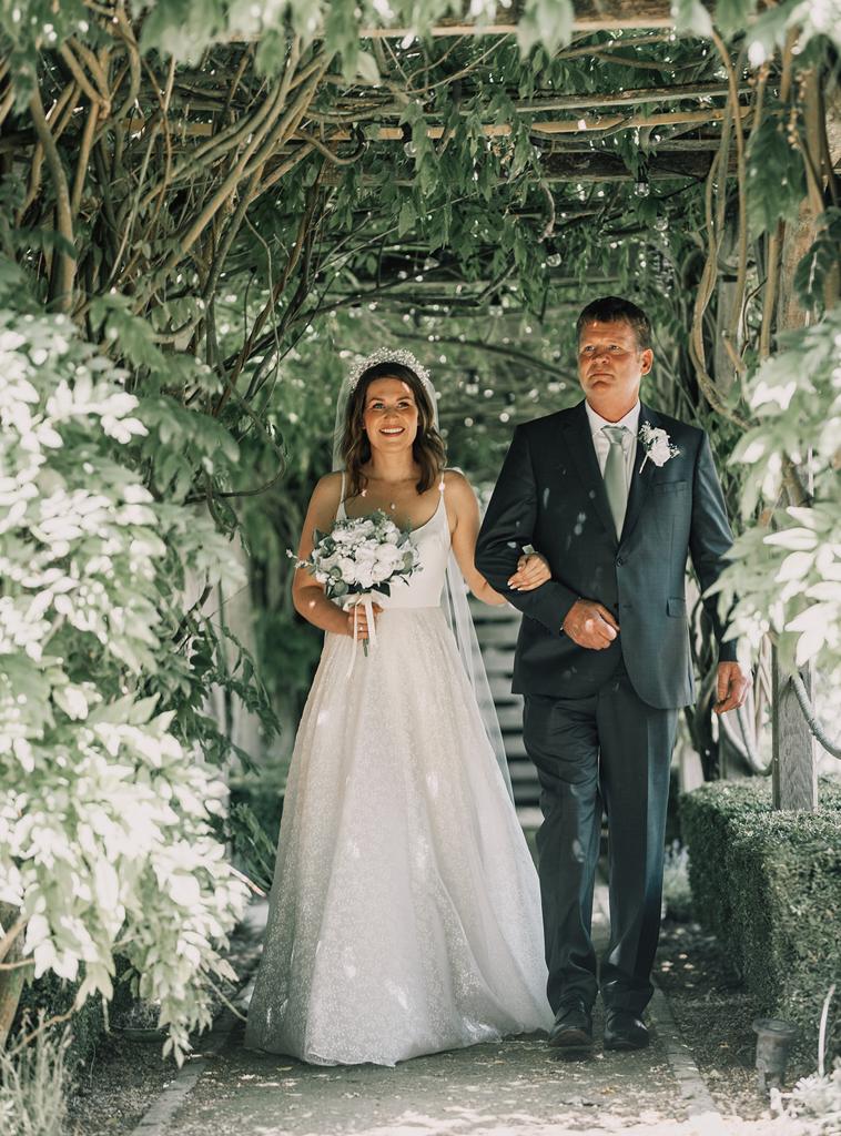 Erin walking down the aisle with her Dad under the arch in the courtyard of The Tythe Barn in Launton near Bicester - photo by The Kensington Photographer