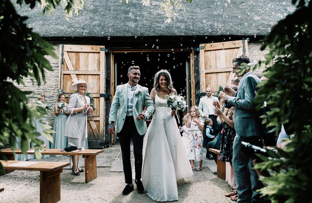 Erin and Vinnie walk back down the aisle of their outdoor ceremony surrounded by bubbles in the courtyard of The Tythe Barn - photo by The Kensington Photographer