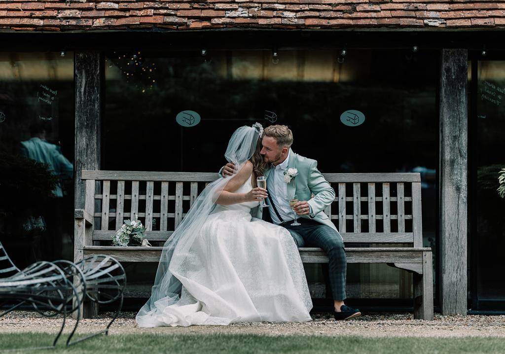 Vinnie and Erin hugging sat on a bench in the courtyard of The Tythe Barn - photo by The Kensington Photographer