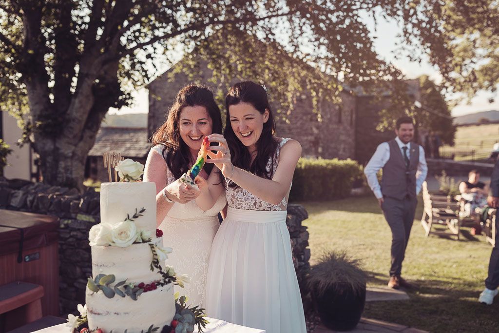 Kay and Rach cutting their wedding cake revealing rainbow coloured layers. Sophisticated Barn Wedding. Photography by Tiree Dawson Photography. Videography by Veiled Productions. Sophisticated New House Farm wedding in the Summer 2019. Cake made by Baba Ganoush Catering York.