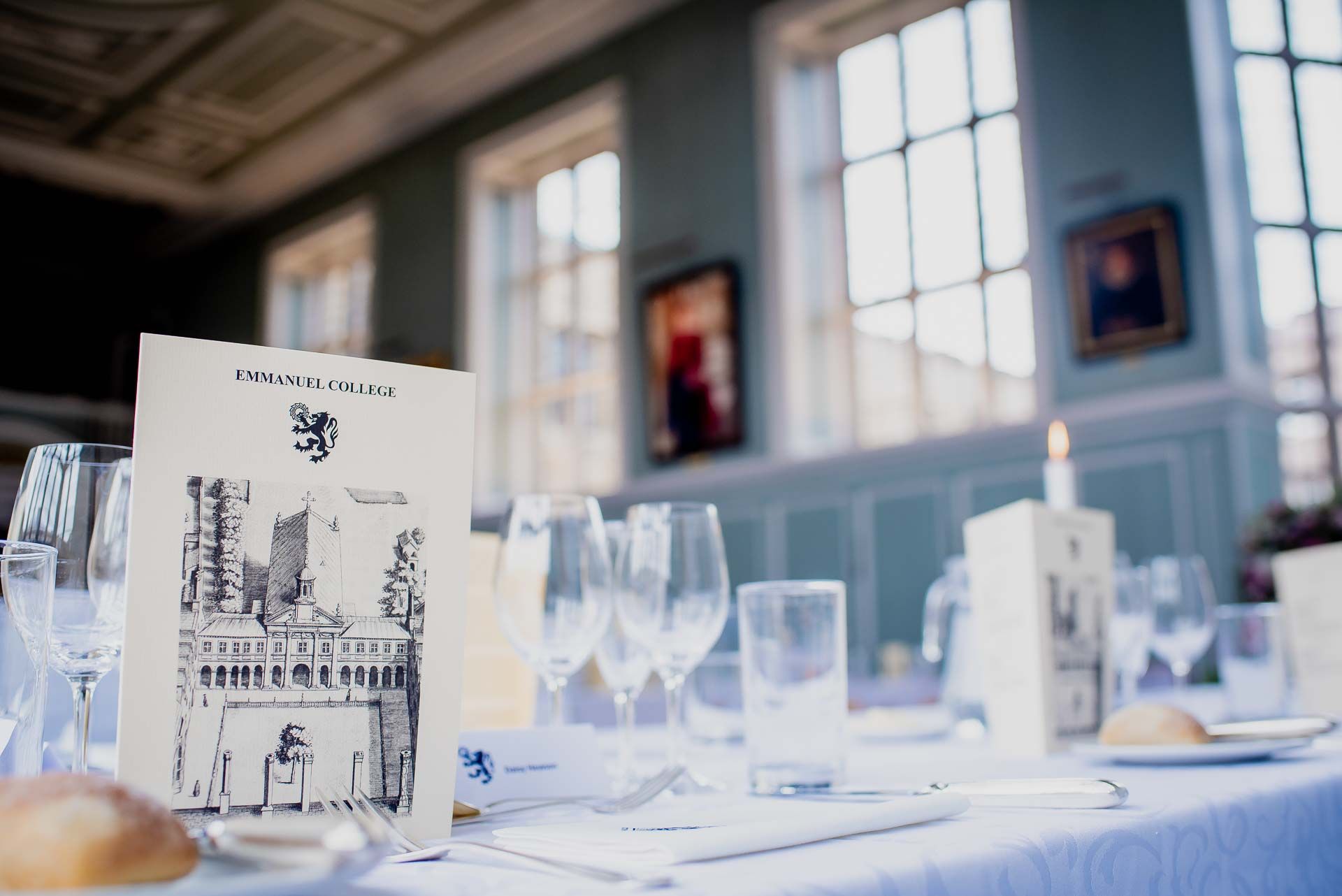 The table set for the wedding breakfast with the menu cover stood upright on the table displaying Emmanuel College image. A candle and glassware is blurred in the background against the big glass windows. Photo thanks to Damien Vickers Photography.