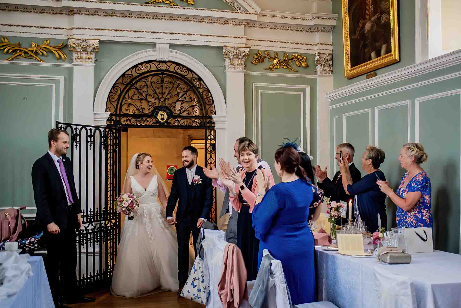 Leah and Nic being announced into the wedding breakfast as newlyweds in the magnificent hall of Emmanuel College Cambridge. Their guests are applauding whilst they are smiling at each other. Photo credit to Damien Vickers Photography.