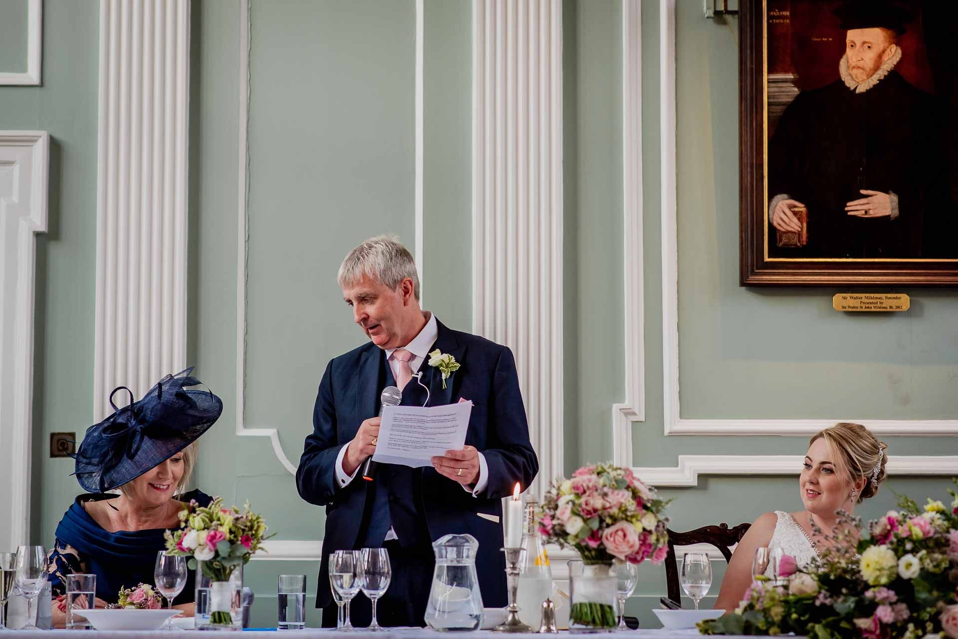 Leah's Dad giving his speech during their wedding breakfast at Emmanuel College Cambridge. Beautiful flowers are sat in vases on the table. Photo thanks to Damien Vickers Photography.