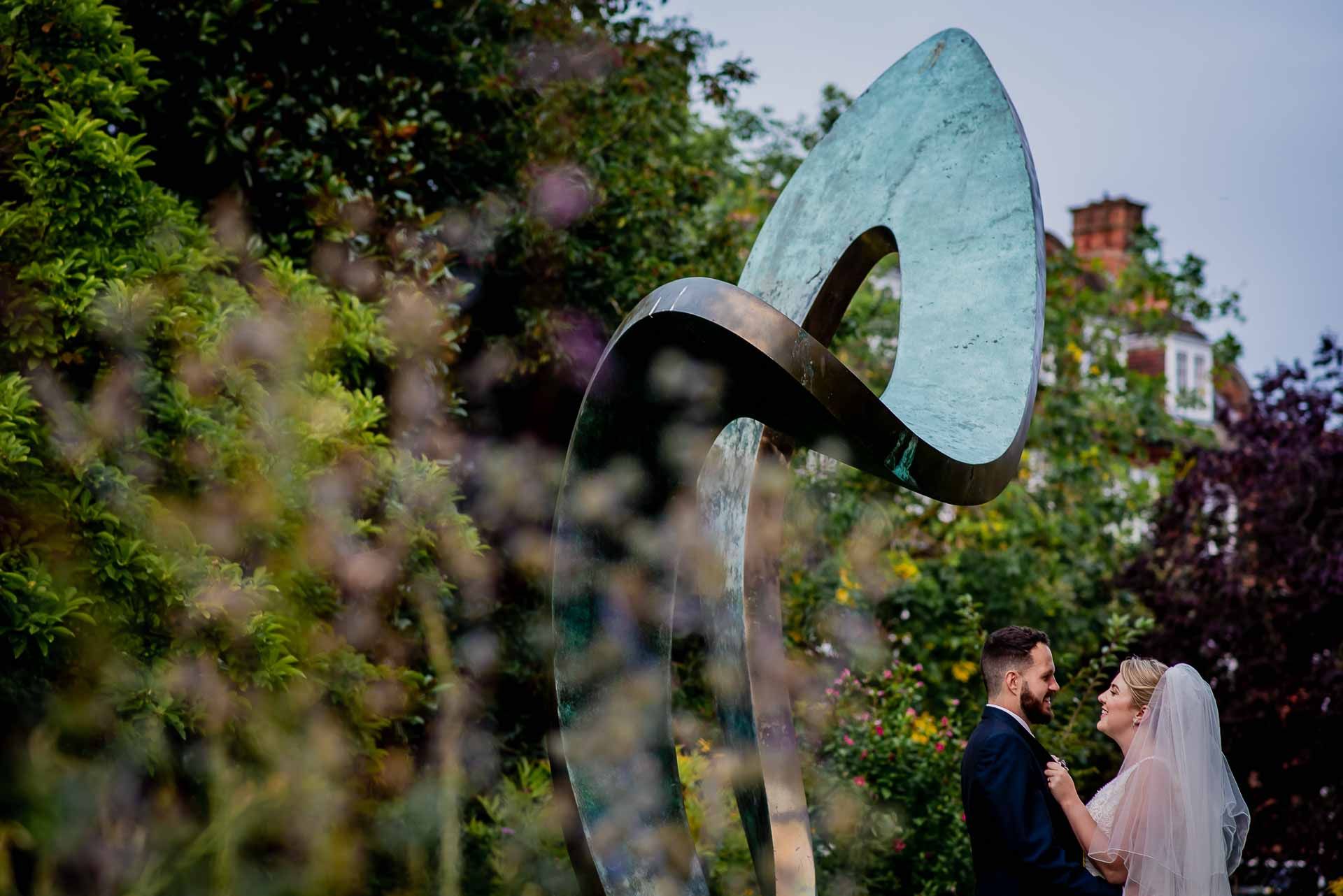 Leah and Nic in the gardens of Emmanuel College, Cambridge University - stood beneath a sculpture with trees surrounding. Photo by Damien Vickers Photography. 