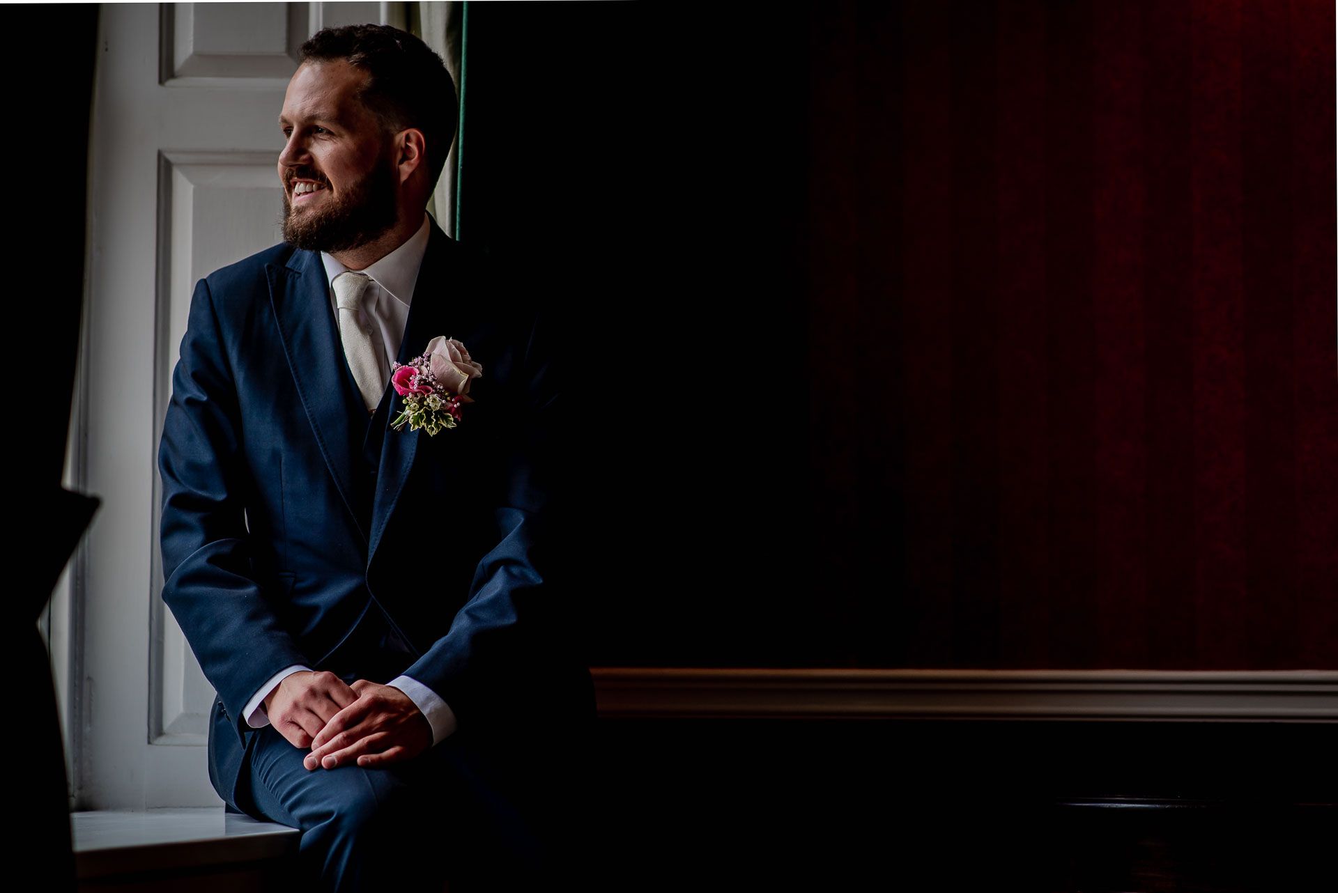 Nic ready to get married to Leah with a navy jacket and pink rose button hole. He is sitting on the windowsill looking out at the grounds of Emmanuel College in Cambridge. Photo by Damien Vickers Photography. 