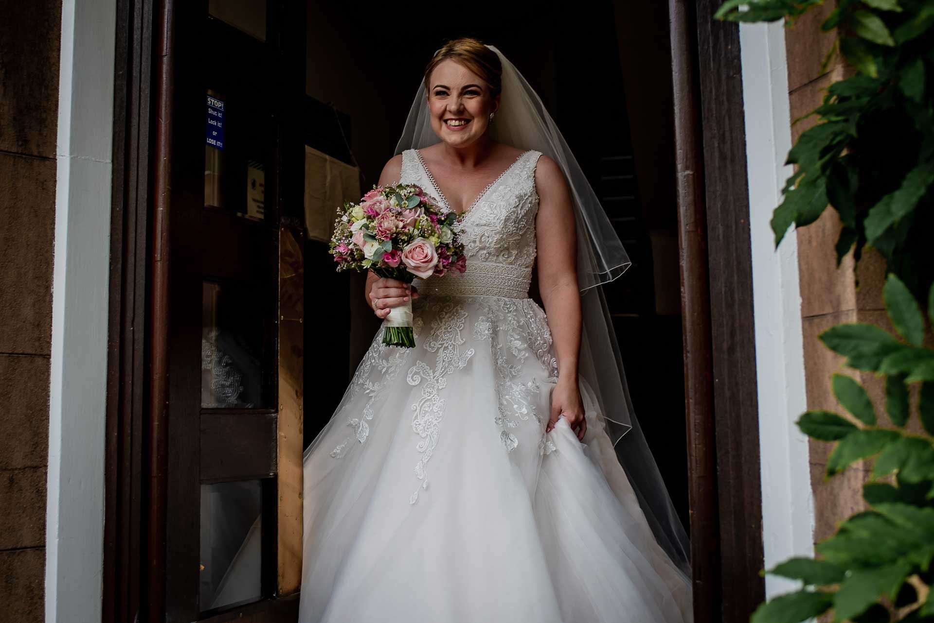 Leah walking through the door into the courtyard of Emmanuel College in her wedding dress and holding her bridal bouquet. Photography by Damien Vickers Photography.