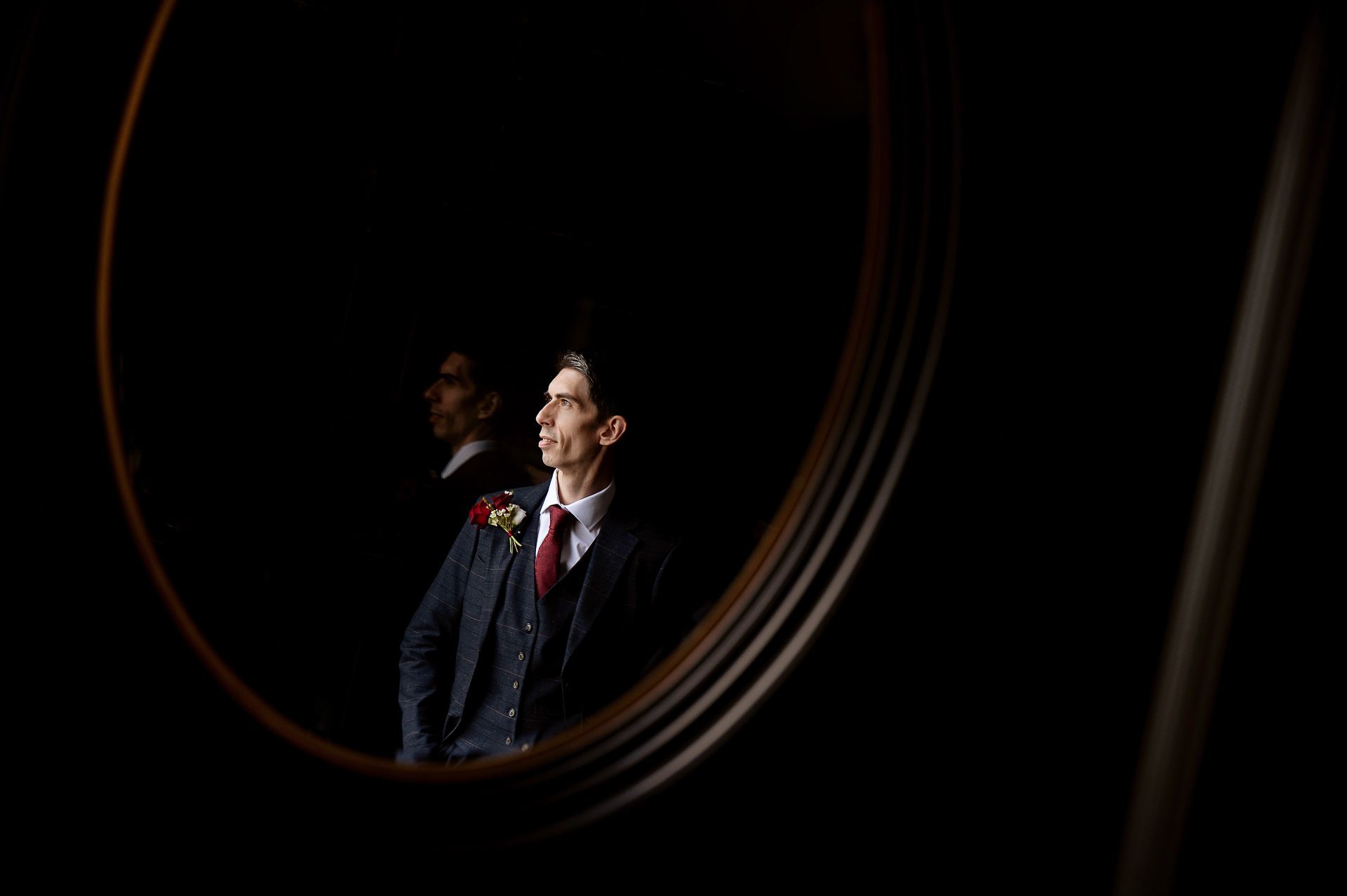 A reflection in a round mirror of Ross in his navy suit with a red tie and red buttonhole flower at Swynford Manor. Photography by Fountain Photography. Videography by Veiled Productions