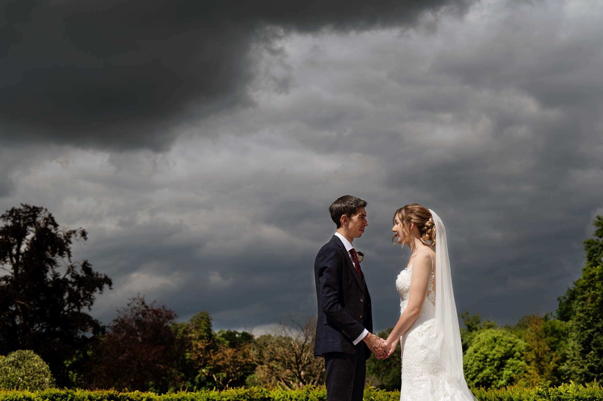 Sophie and Ross holding hands in the gardens of Swynford Manor with dark, moody skies behind them. Photography by Fountain Photography. Videography by Veiled Productions.