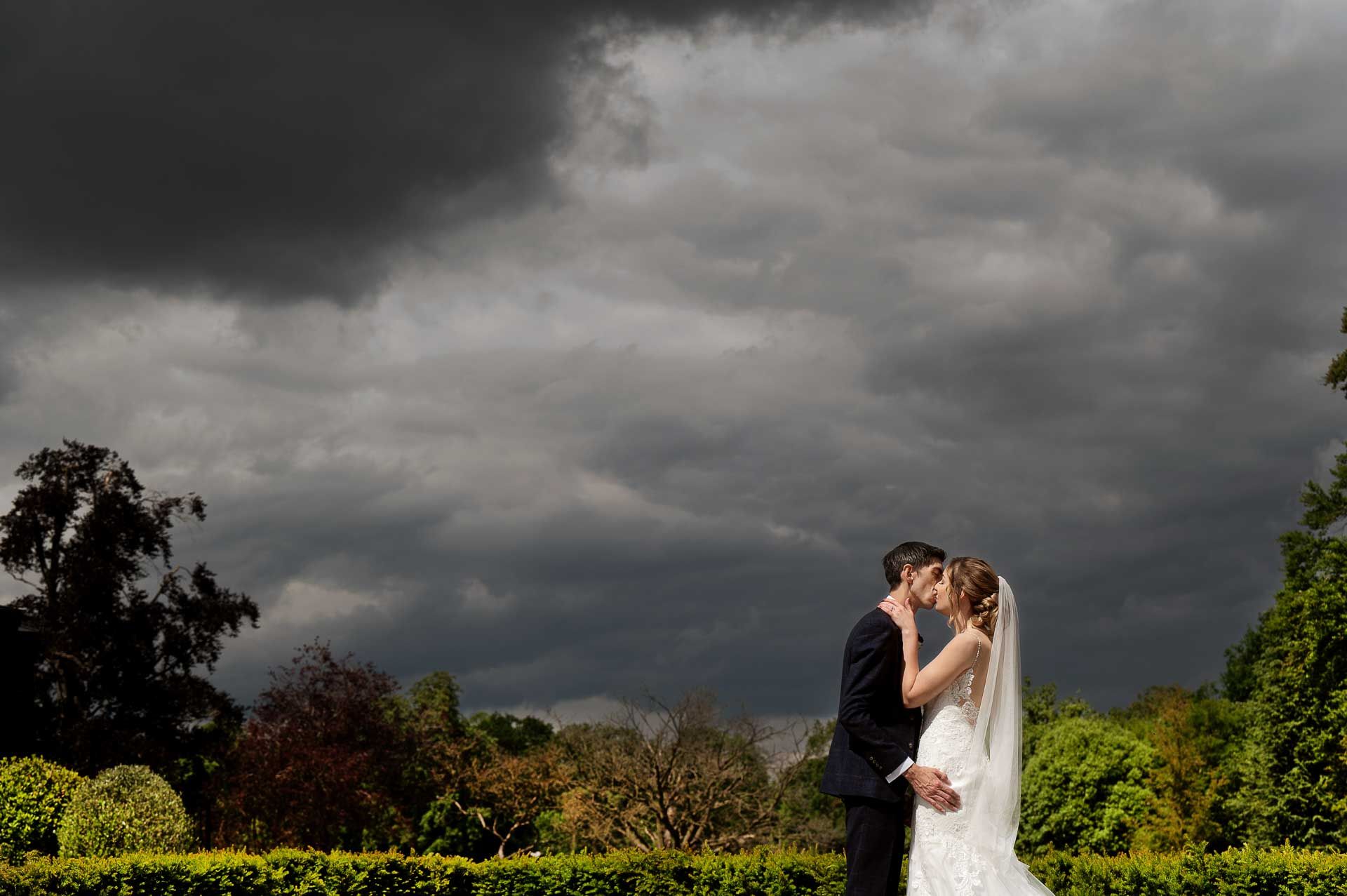 Sophie and Ross kissing in the gardens of Swynford Manor with dark, moody skies behind them. Photography by Fountain Photography. Videography by Veiled Productions.