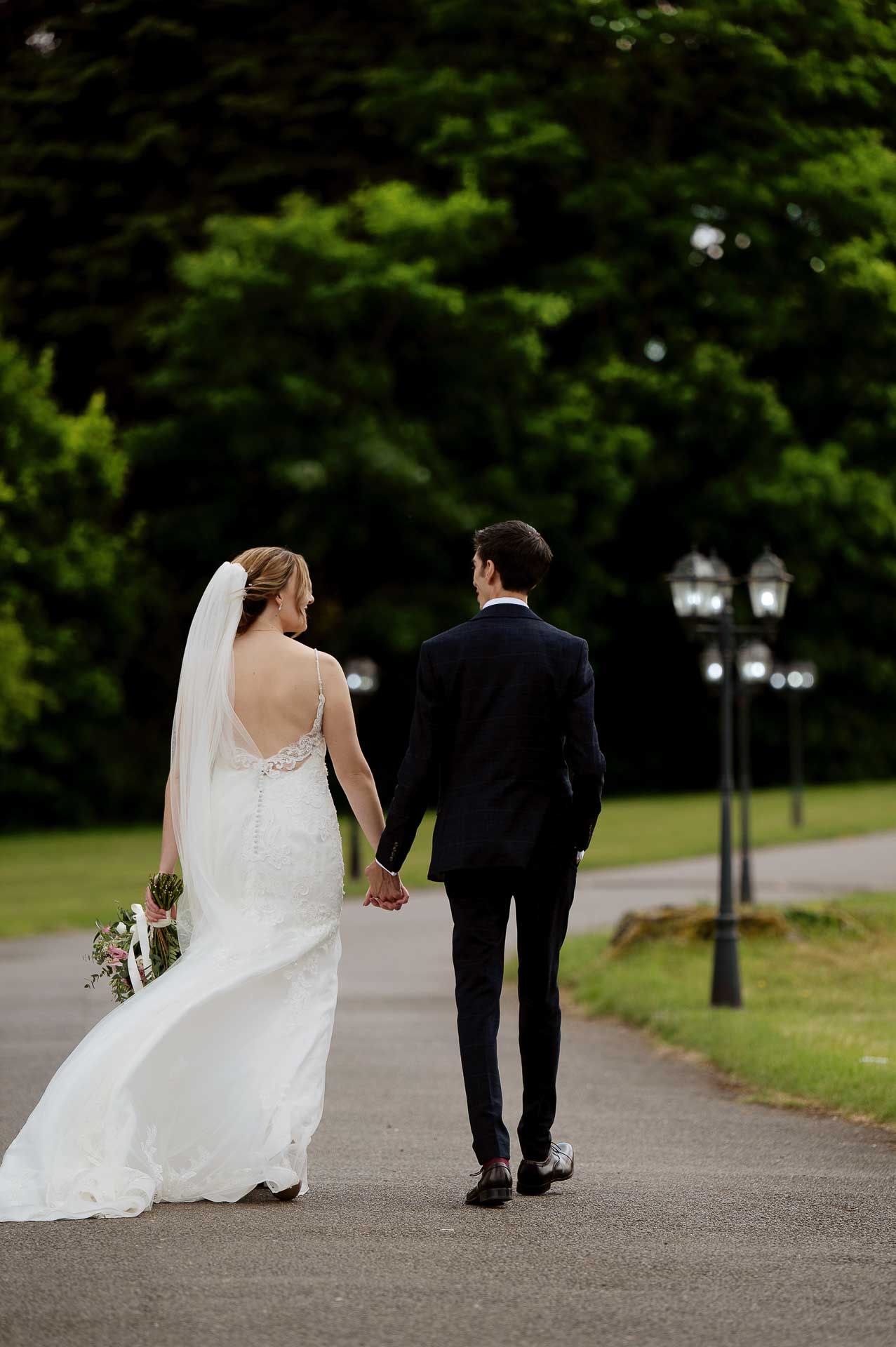 Sophie and Ross holding hands walking away from the camera down the driveway at Swynford Manor. Photography by Fountain Photography. Videography by Veiled Productions.