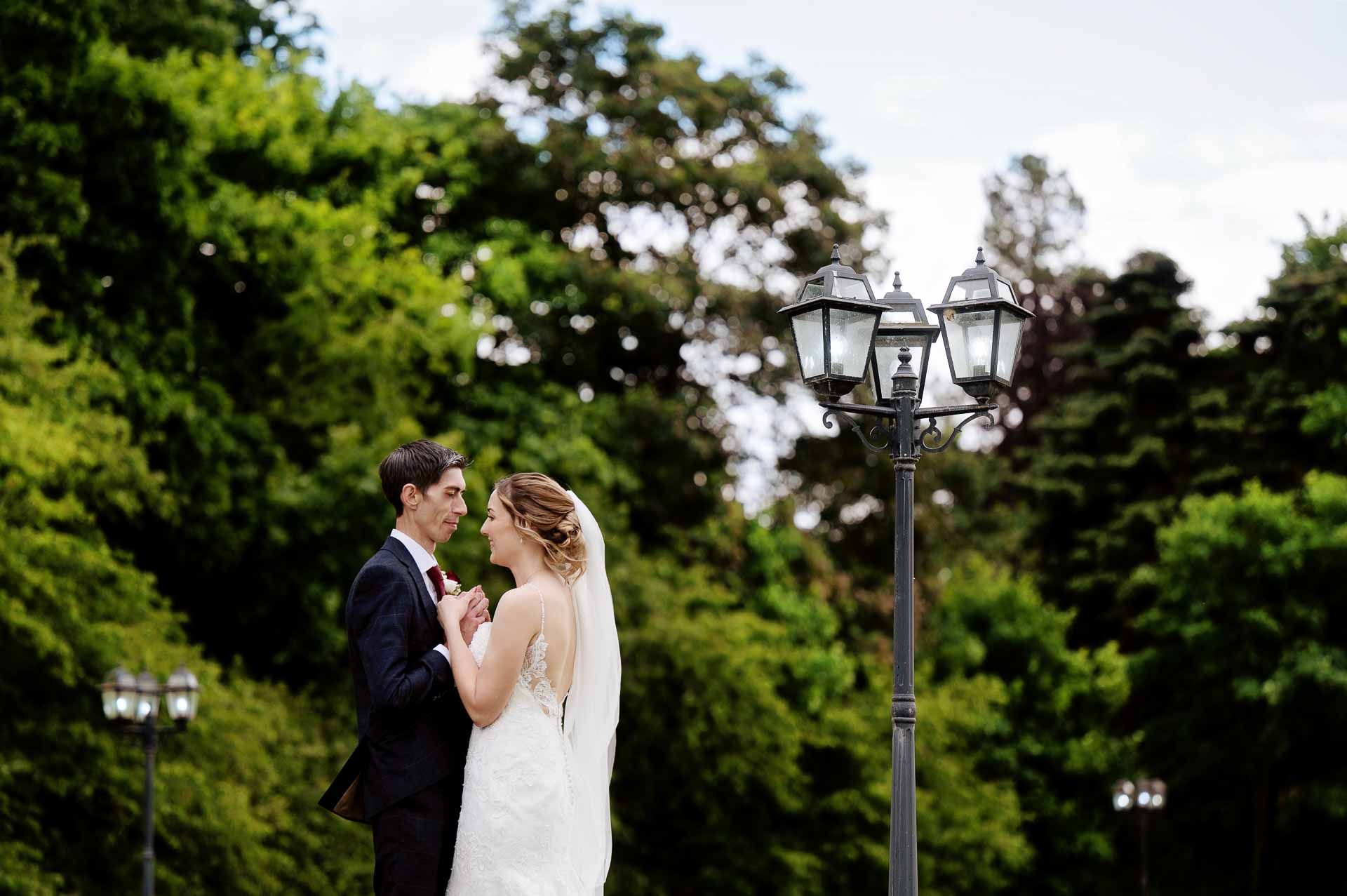 Sophie and Ross holding hands, smiling at each other stood between two lamp posts during their couples photoshoot at Swynford Manor. Photography by Fountain Photography. Videography by Veiled Productions.