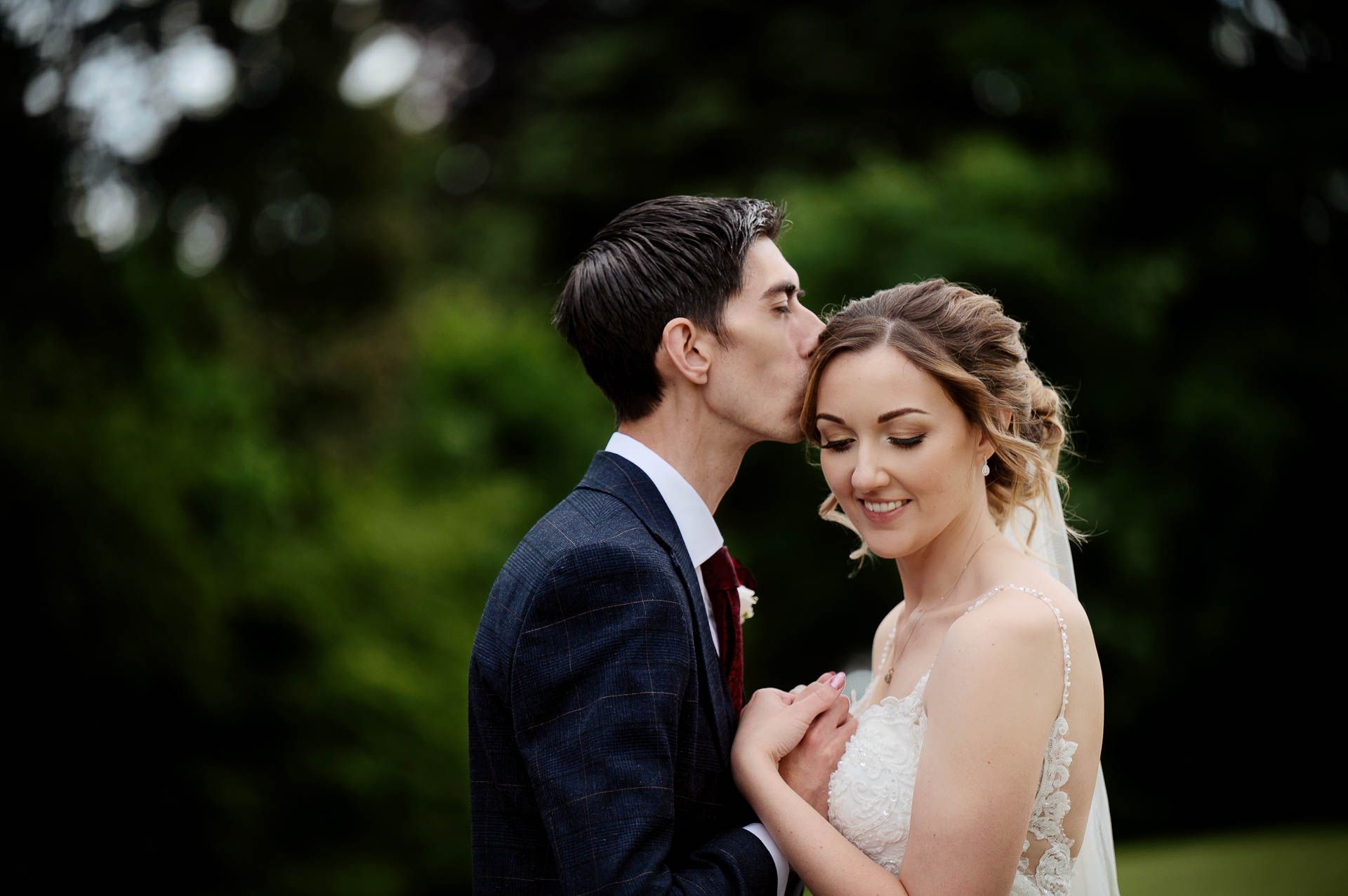 Sophie and Ross holding hands with Ross kissing Sophie's head and Sophie smiling, looking at the floor during their couples photoshoot at Swynford Manor. Photography by Fountain Photography. Videography by Veiled Productions.