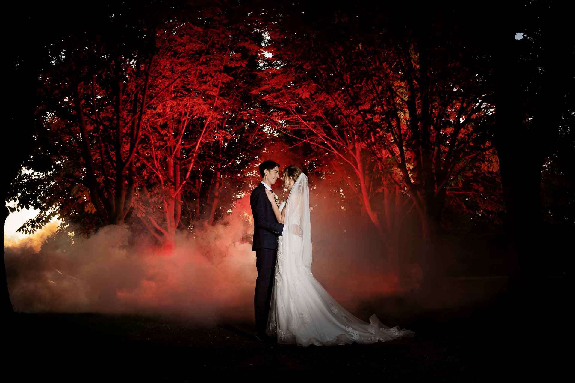 A wide photo amongst trees with a smoke bomb and red lighting circulating Ross and Sophie during sunset at Swynford Manor. Photography by Fountain Photography. Videography by Veiled Productions.