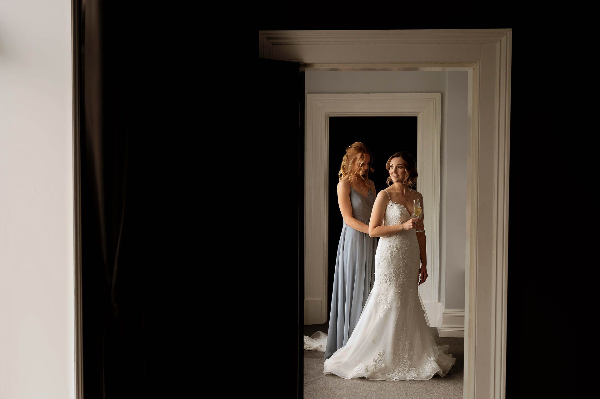 Sophie's bridesmaid helping secure Sophie's wedding dress. The two of them are stood in the doorway of the wedding suite at Swynford Manor, Sophie is holding a glass of Champagne and looking across the room. Photography by Fountain Photography. Videography by Veiled Productions.