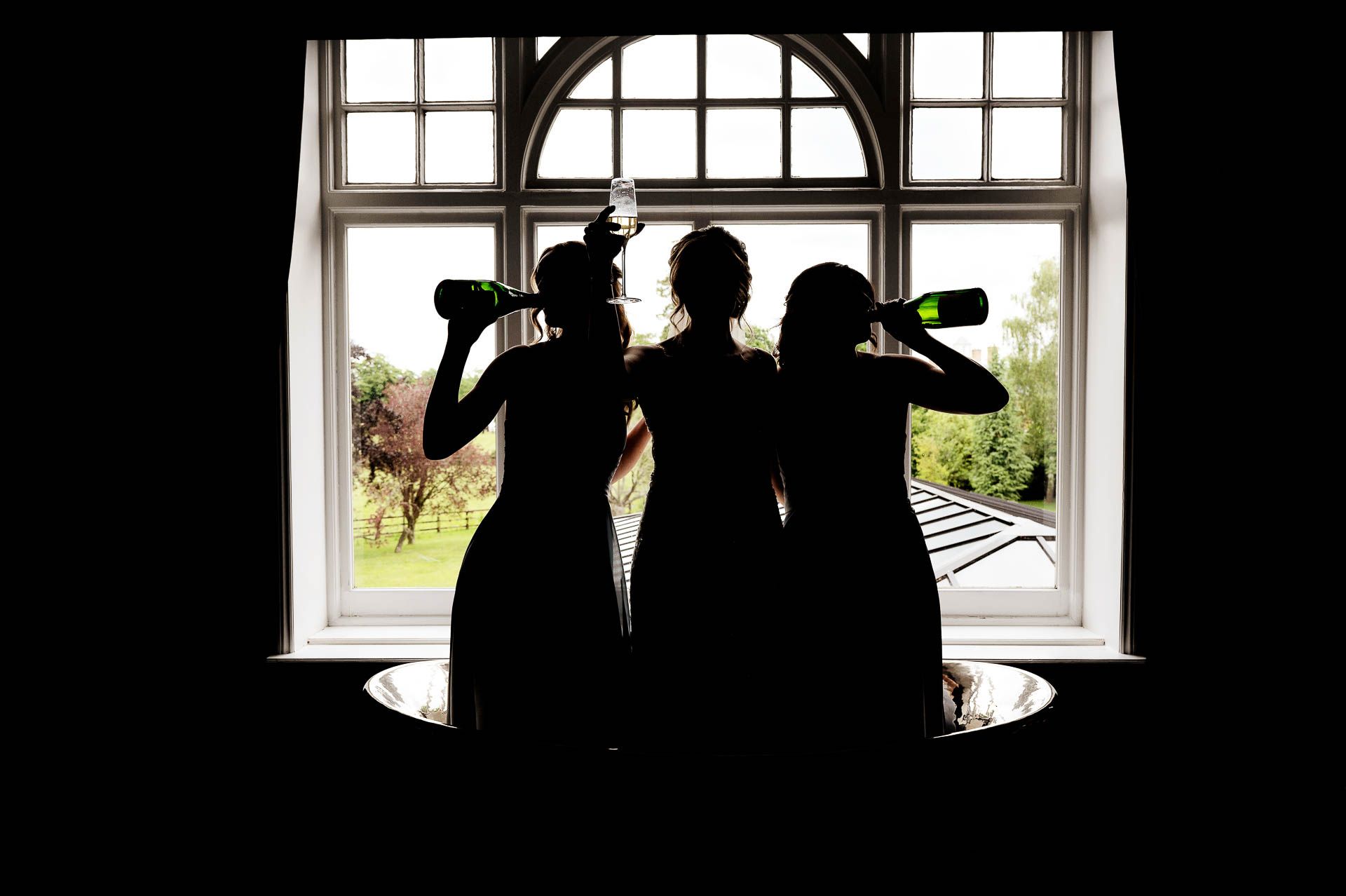 Sophie and her bridesmaids silhouetted in the window of the honeymoon suite toasting with champagne at Swynford Manor. Photography by Fountain Photography. Videography by Veiled Productions.