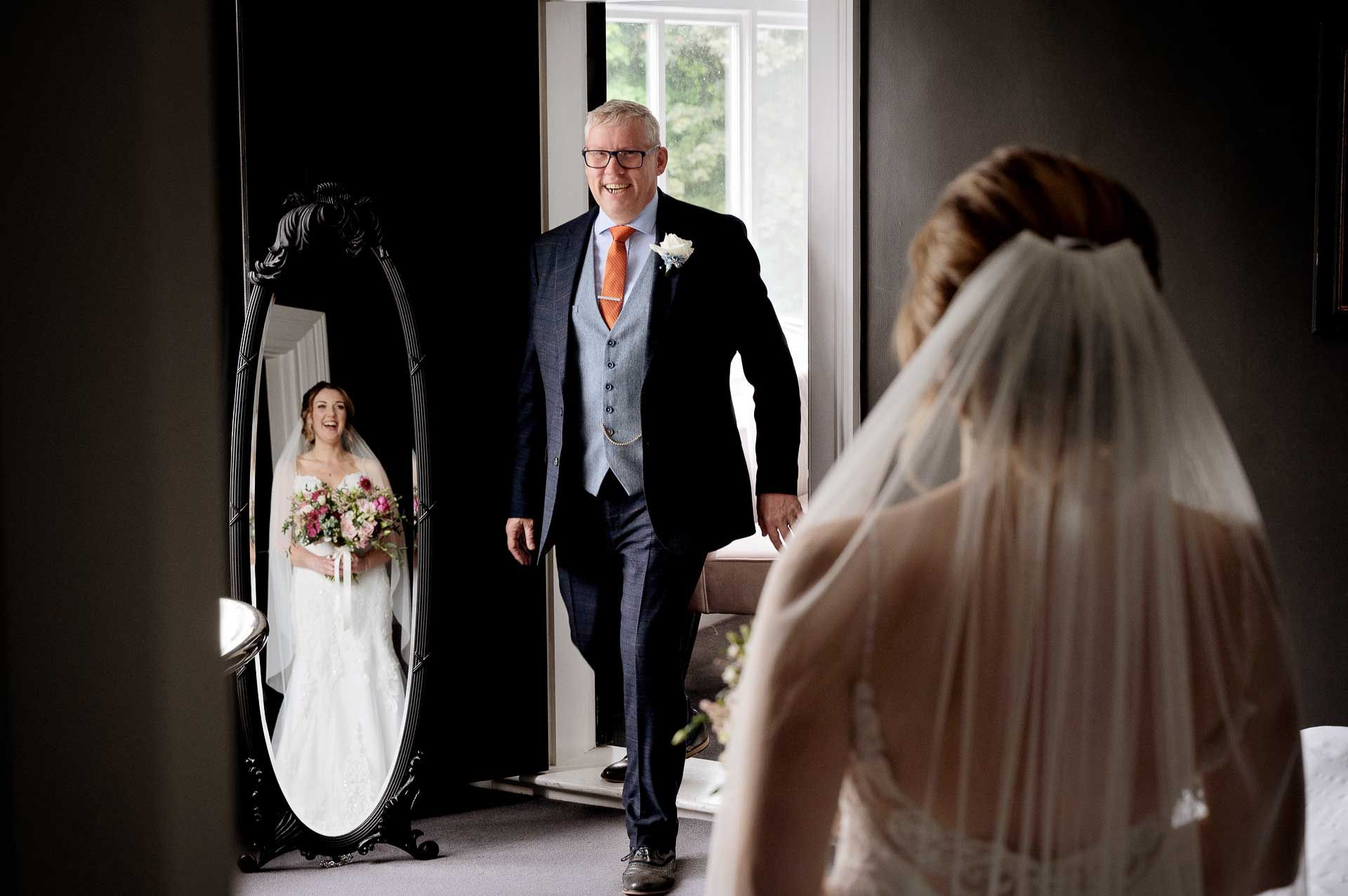 Sophie's Dad seeing her in her wedding dress for the first time at Swynford Manor. Sophie's reflection can be seen in the mirror, they are both grinning. Photography by Fountain Photography. Videography by Veiled Productions.
