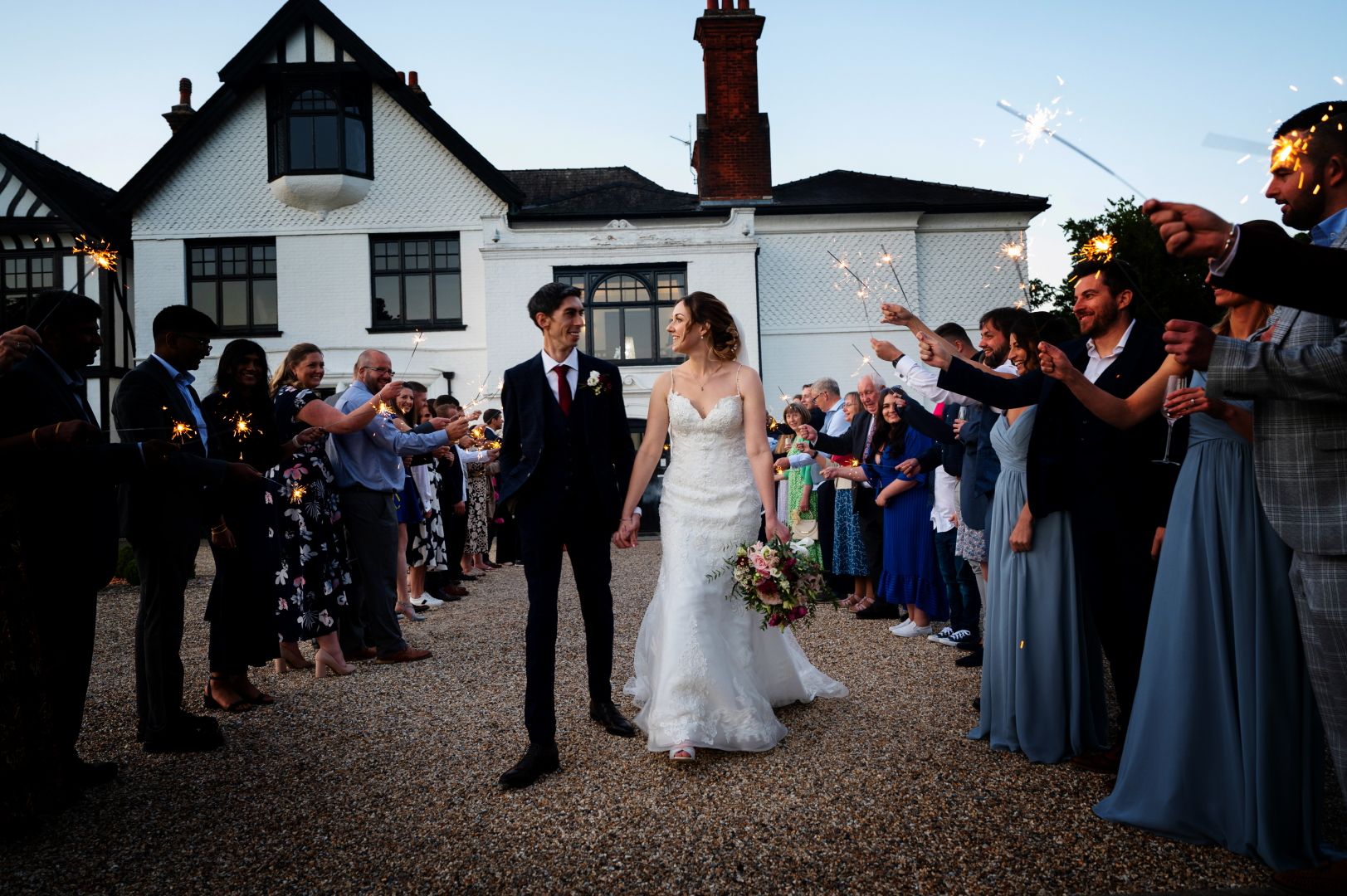 Sophie and Ross grinning during their sparkler aisle at dusk outside Swynford Manor. Photo by Fountain Photography.