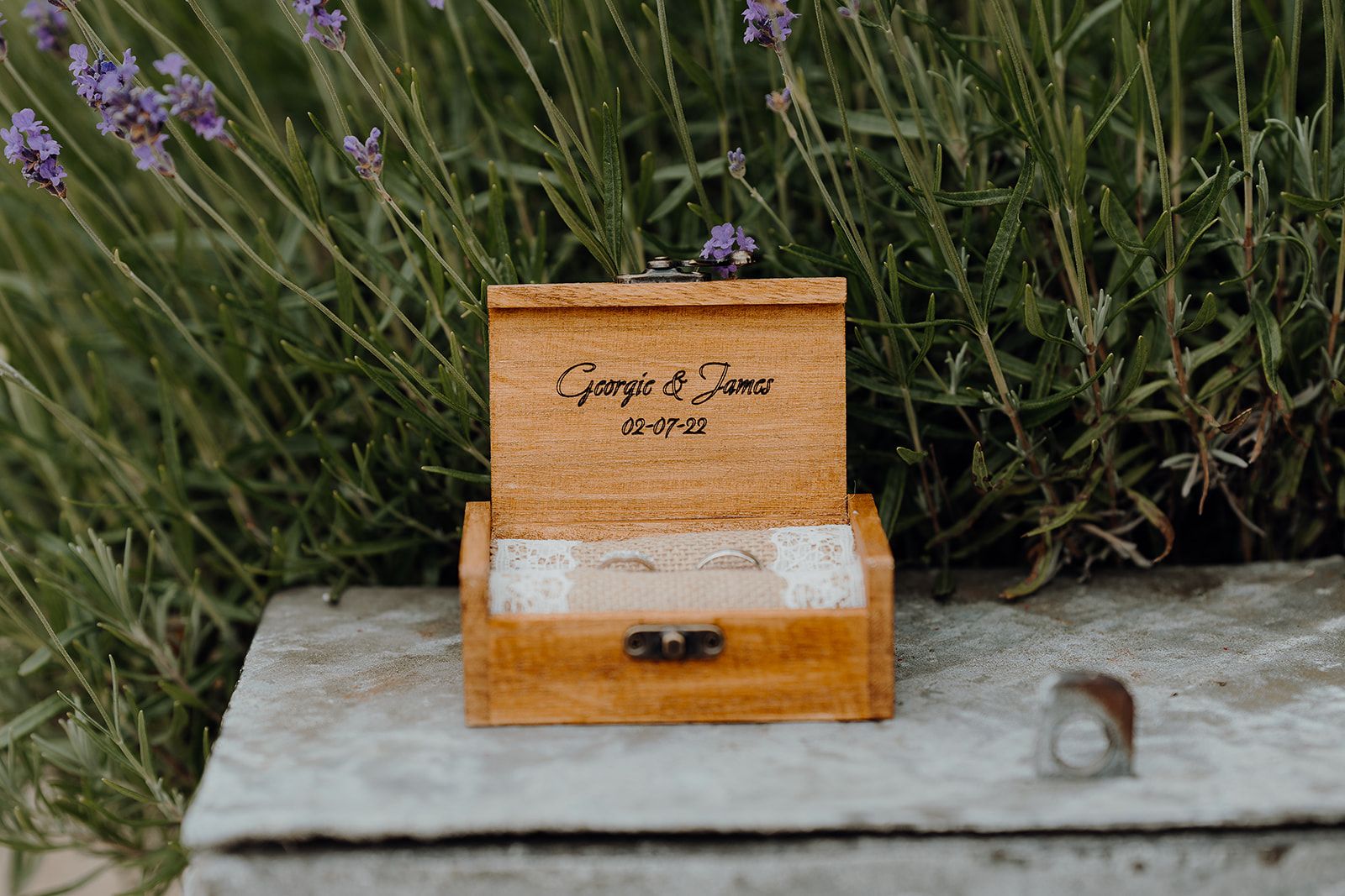 Personalised wooden wedding ring with Georgie and James names plus the date of their wedding and a hessian insert. Lavender flowers in the background. Photo thanks to Sam and Steve Photography.