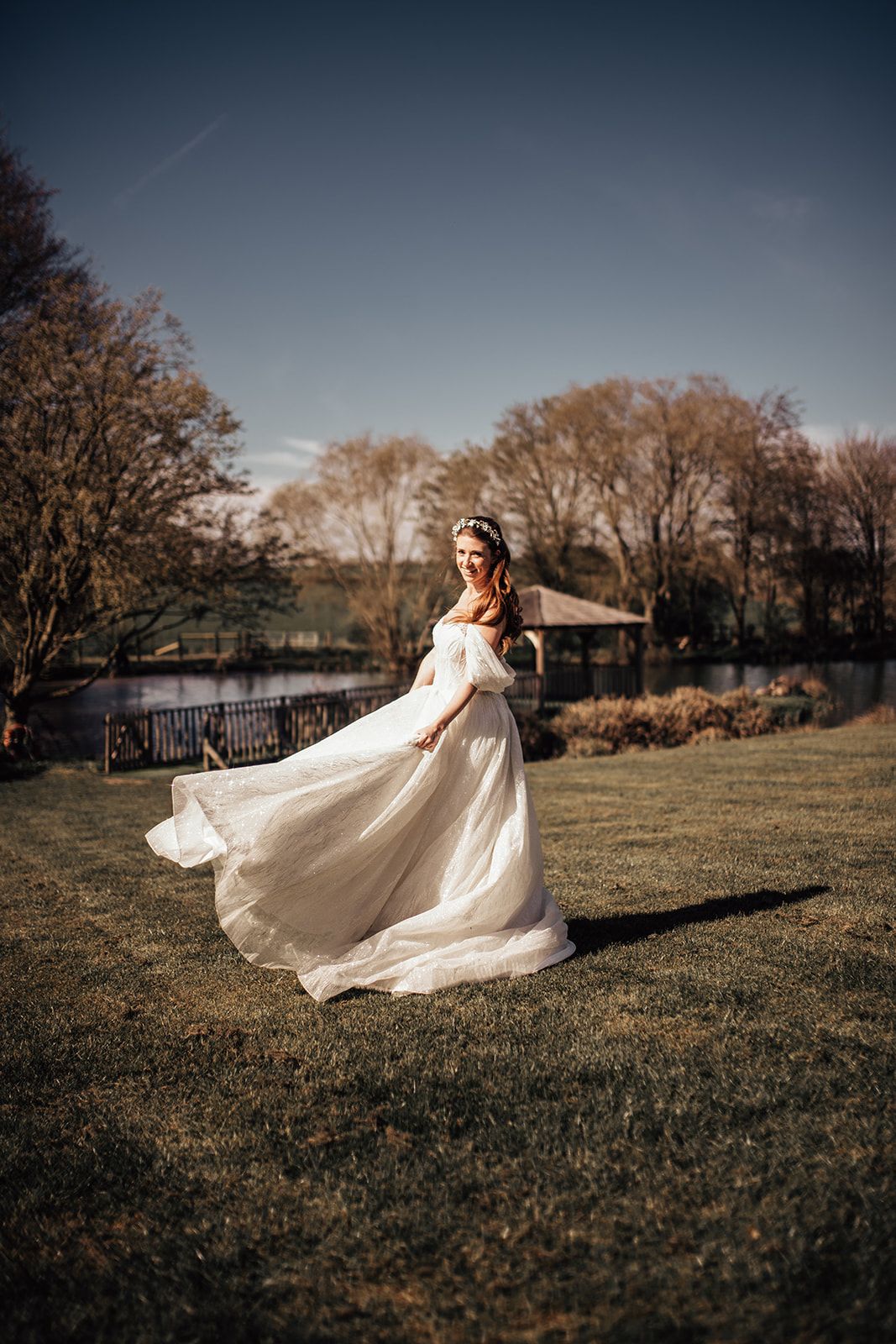 A bride stood on the lawn in front of the lake and gazebo at Furtho Manor Farm sweeping her dress and looking over her shoulder. Photo thanks to Francesca Checkley Photography.