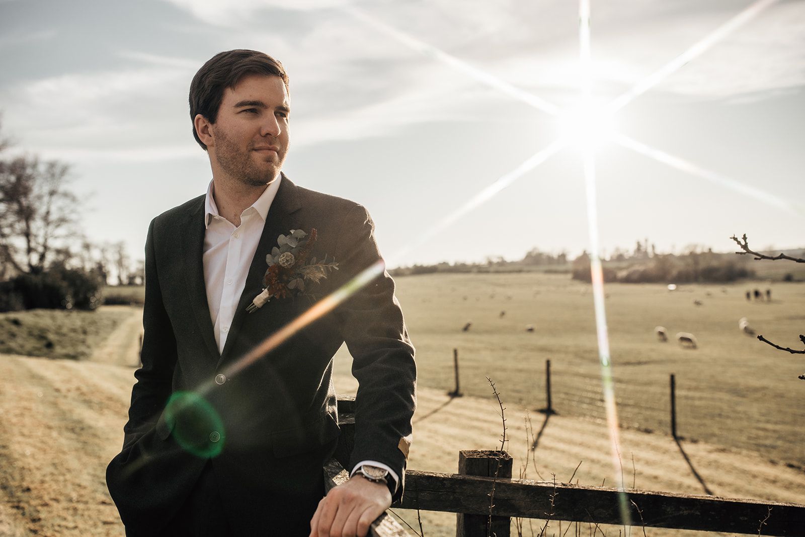 Photo of a groom leaning against a fence with an open collar and gorgeous floral button hole. The countryside and sheep are in the background with the suns rays reflecting across the image. Photo thanks to Francesca Checkley Photography.