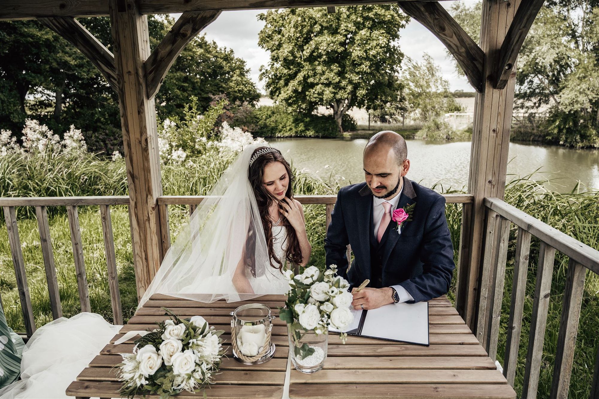 Newlyweds signing the register under the gazebo on the lake at Furtho Manor Farm. The lake and trees are in the background. Photo thanks to Francesca Checkley Photography.