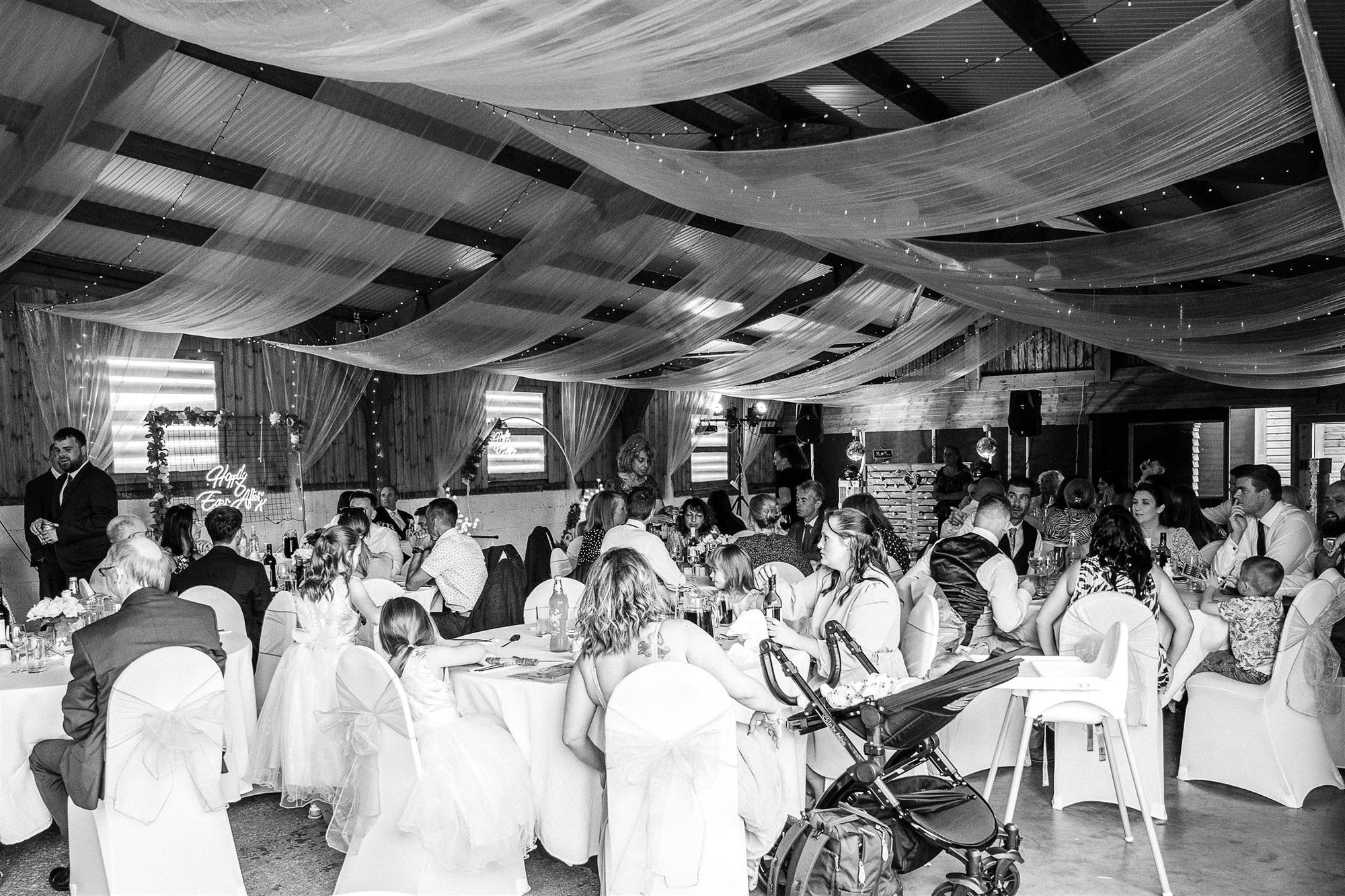 Wide angle view during a wedding breakfast of the barn at Furtho Manor Farm. Drapes and fairy lights decorate the ceiling which give it a cosy feel. Photo thanks to Francesca Checkley Photography.