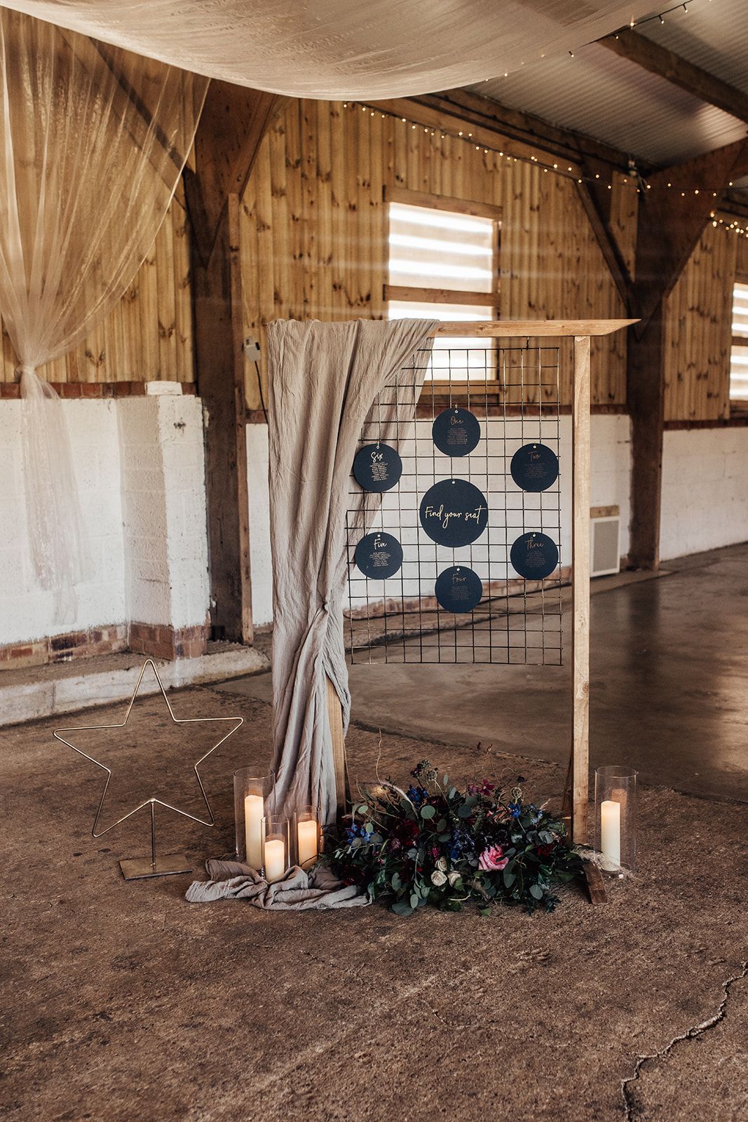 Beautiful table plan with charcoal circles representing the tables attached to a grid. A star, candles and floral arrangement sit underneath the frame and some fabric hangs on the left hand side of the frame giving it an elegant, eye catching look. Photo thanks to Francesca Checkley Photography.