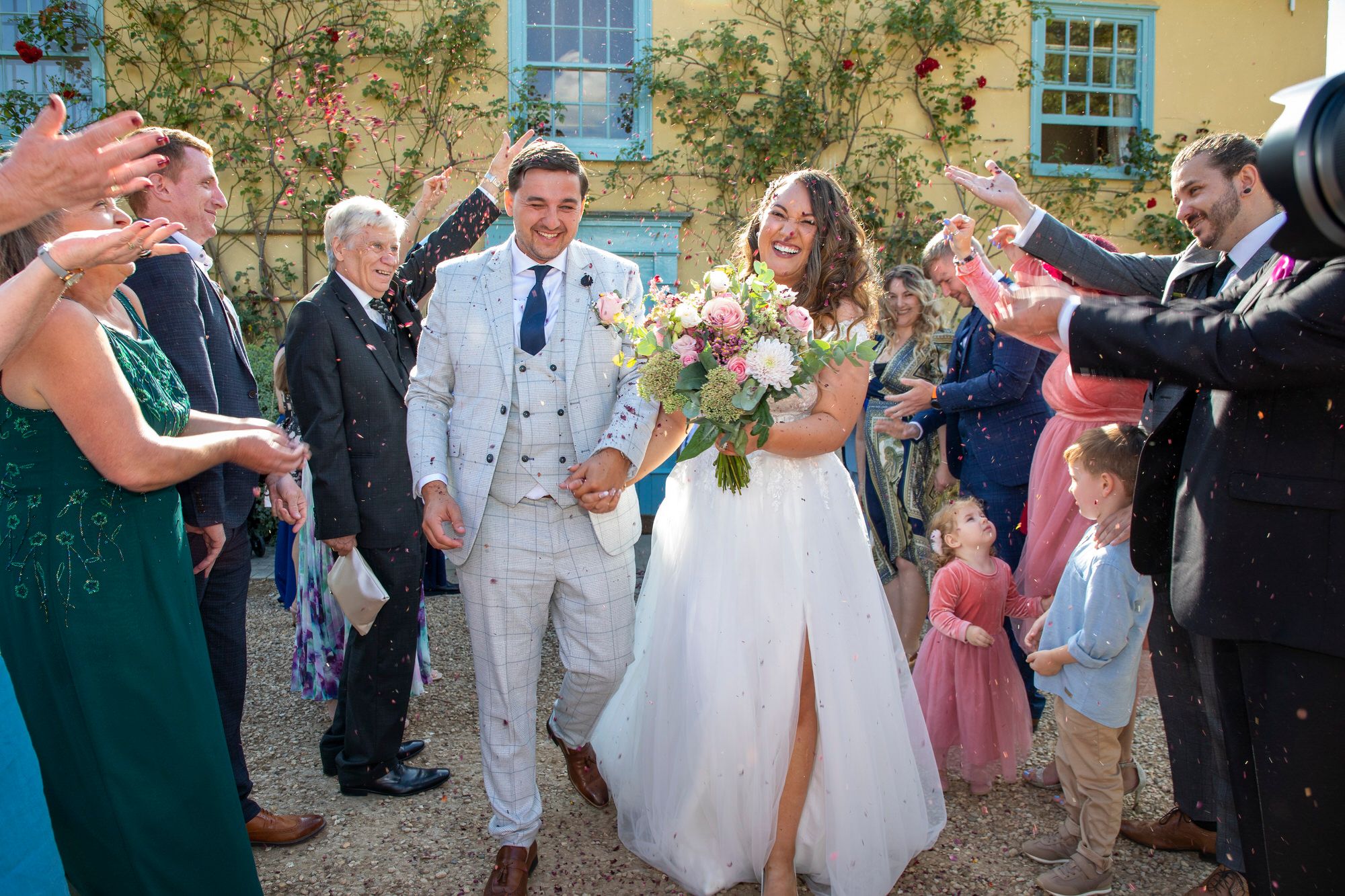Epic confetti photo in front of South Farm's iconic blue and yellow farmhouse. Joanna and Rafal smiling as they walk through the confetti line. Joanna is holding her beautiful pink and green bouquet. Photo thanks to Nigel Charman Photography.