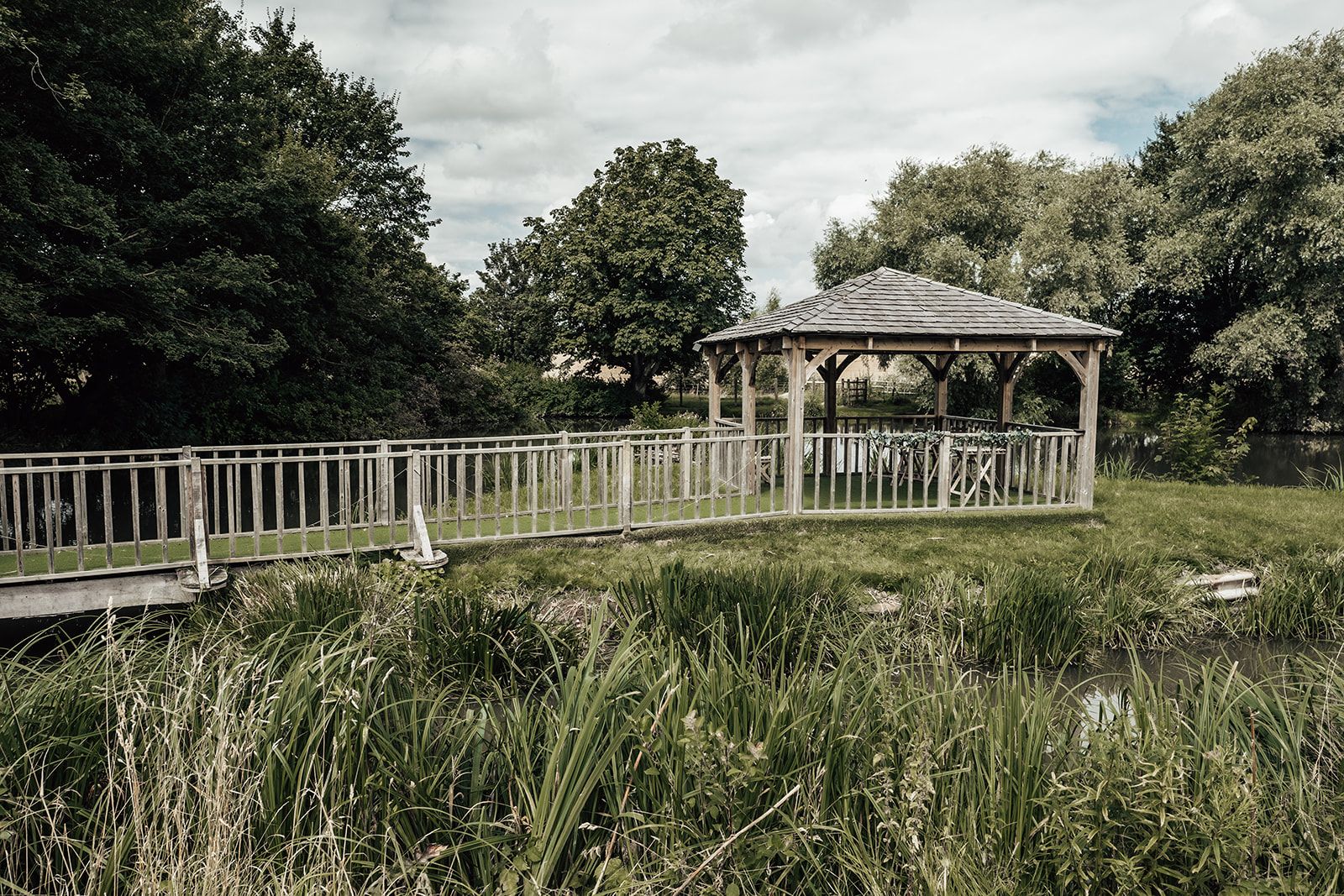 Wide angle landscape view of the gazebo on the lake surrounded by trees at Furtho Manor Farm. Photo thanks to Francesca Checkley Photography.
