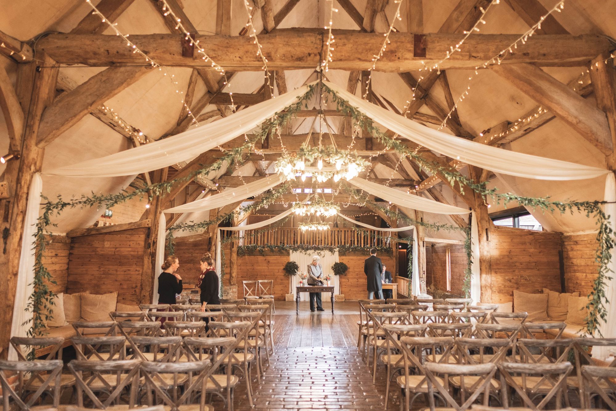 A view from the back of the barn with 3 chandeliers and white drapes hanging from the ceiling at Lains Barn. Beautiful wooden chairs face the front with the wedding coordinators, registrars and James - the groom - stood at the front. Photo thanks to The Falkenburgs via Big Day Productions