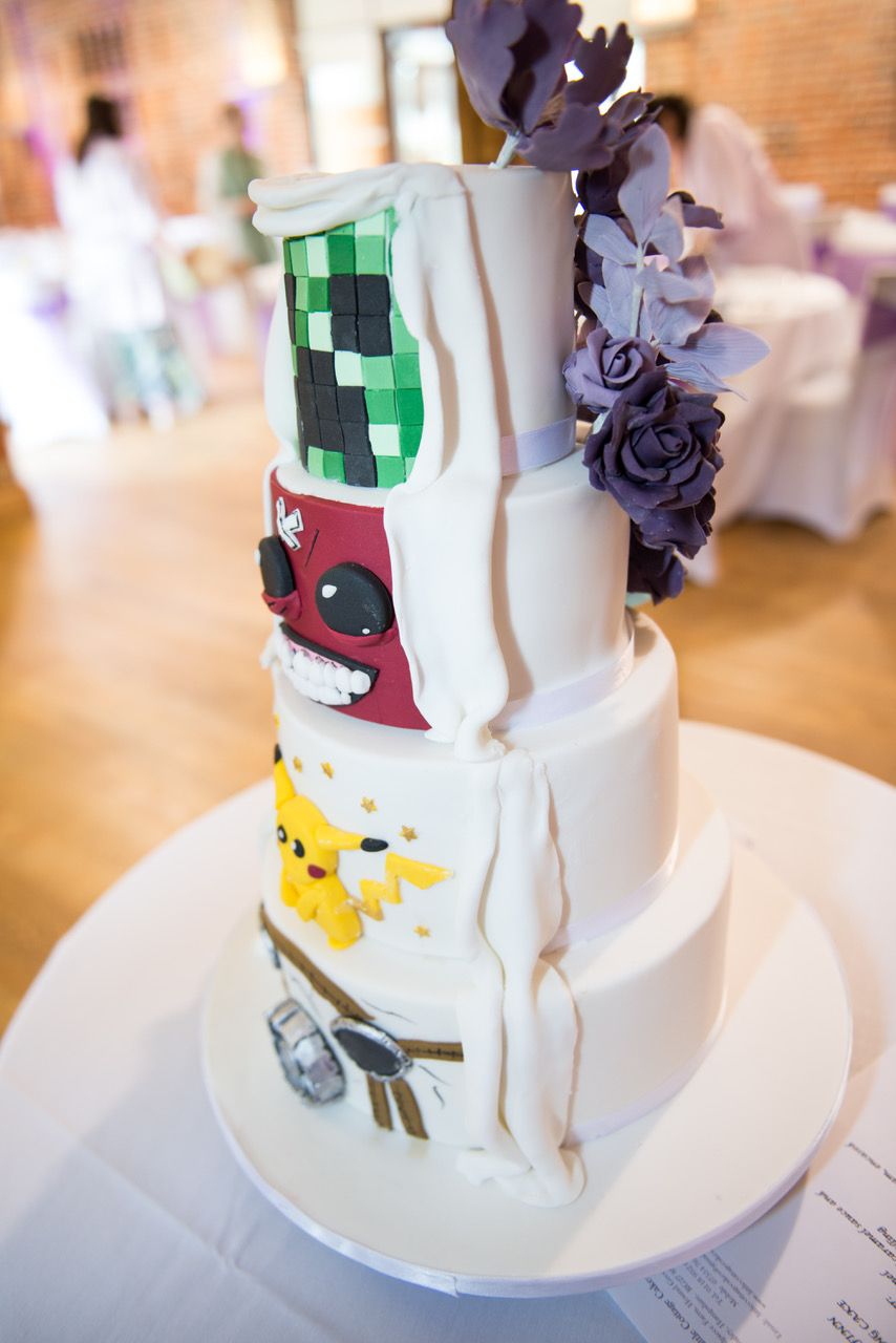 A four tier wedding cake with gaming characters made out of icing on one side and purple icing flowers decorate the other side. Photo thanks to Zoe Warboys Photography at Wasing Park.