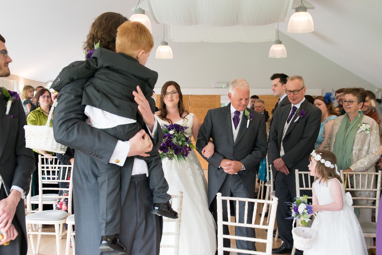 Kelly arriving at the top of the aisle with her Dad to meet Finn who is holding their son whilst their daughter and guests look on. Photo thanks to Zoe Warboys Photography at Wasing Park.