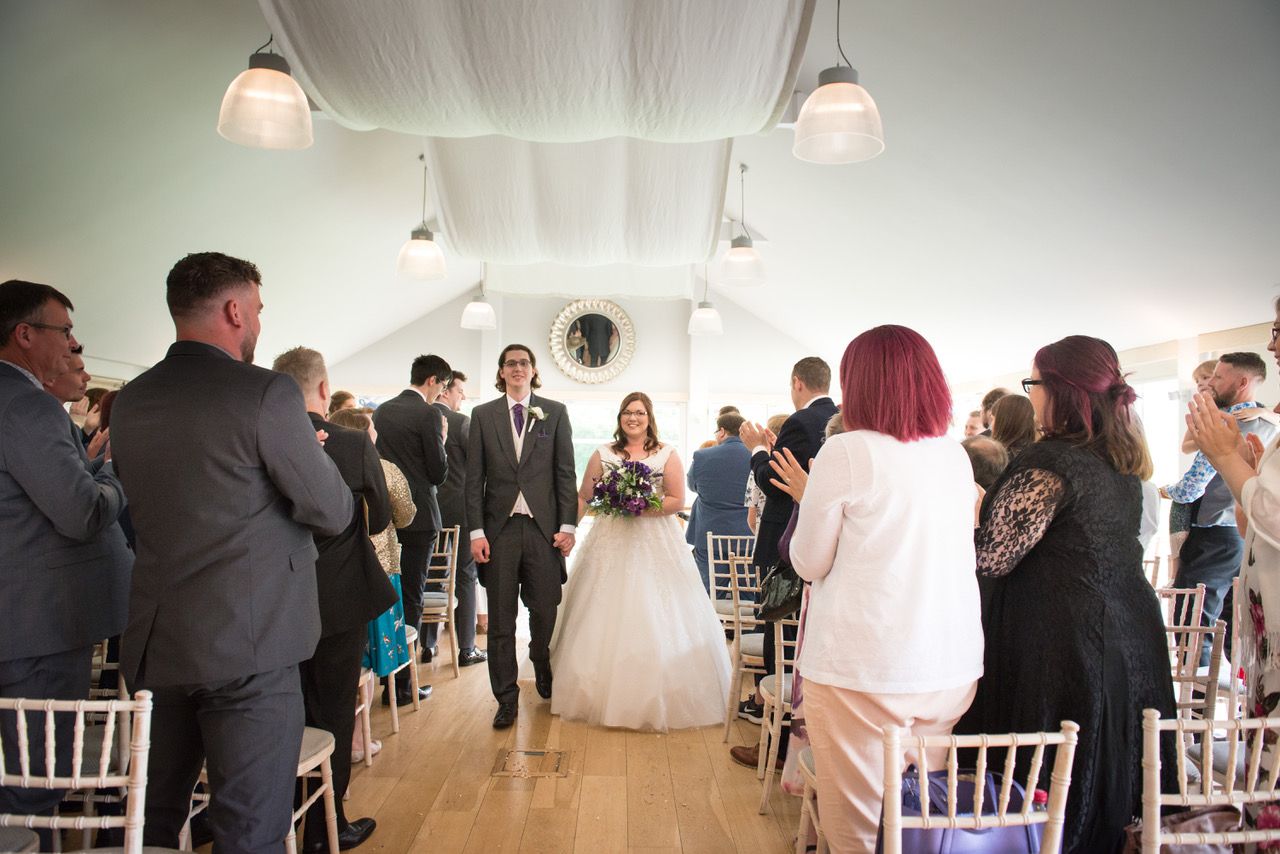 Kelly and Finn exit their wedding ceremony as husband and wife, walking hand in hand and grinning. Kelly is holding her beautiful purple and white bouquet. Photo thanks to Zoe Warboys Photography at Wasing Park.