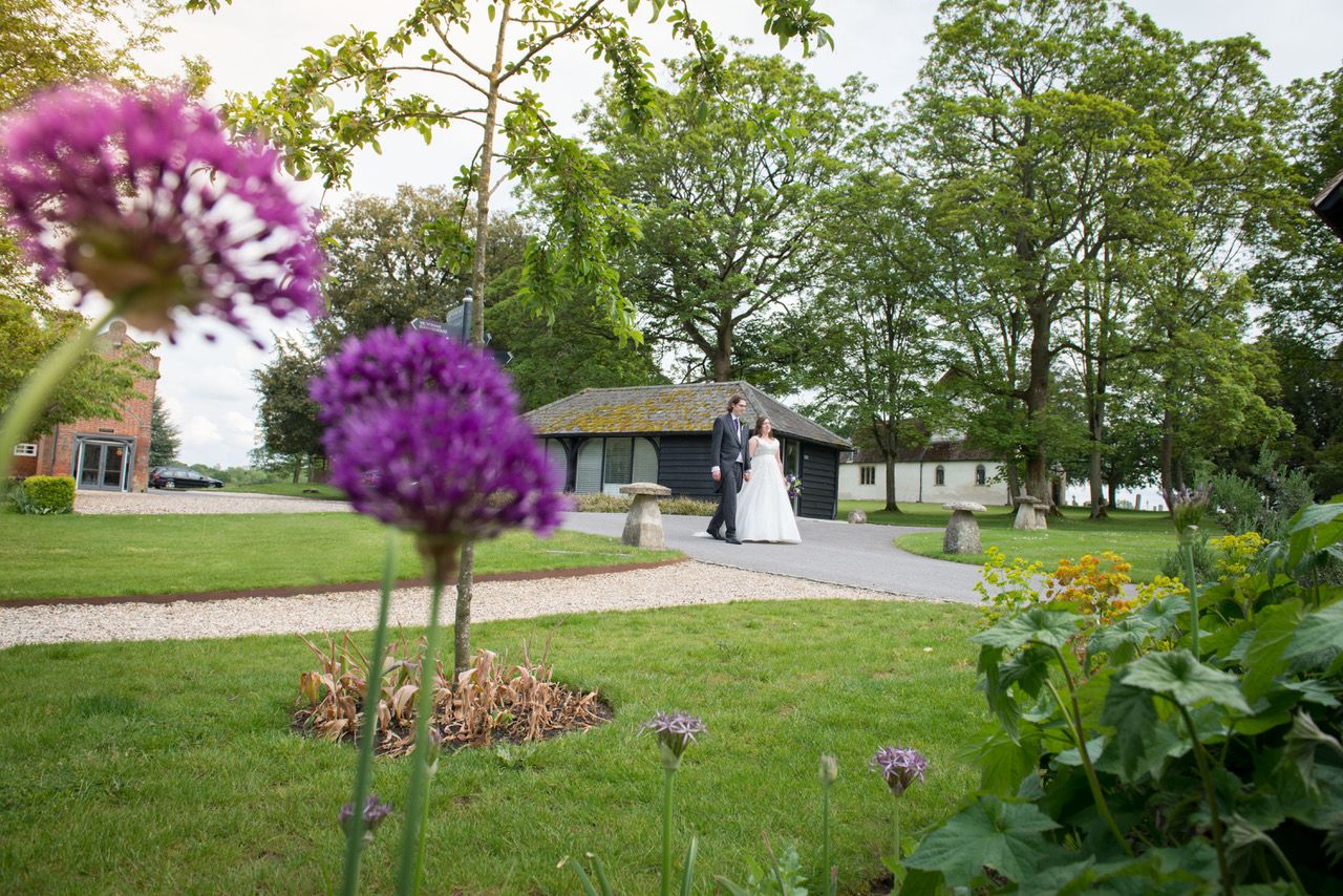 A wide angle photo taken in the gardens of Wasing Park. In the foreground are purple flowers, slightly out of focus and in the background are Kelly and Finn walking hand in hand down the drive at Wasing Park. Photo thanks to Zoe Warboys Photography.
